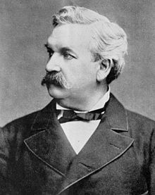 A monochrome photograph of a man from the chest up, approximately 50 years old, with a large moustache and no sideburns, a full head of white hair, looking sharply to the left