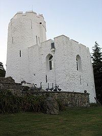 white-painted stone building comprising a square tower and a taller round tower beyond, both with castellations, and a grey stone wall and grass in the foreground