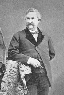 A monochrome photograph of a man from the thigh up, approximately 40 years old, with a large moustache and no sideburns, leaning with his right elbow on a fur-draped arm of furniture, looking slightly to the left