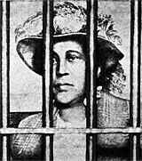 Woman wearing hat staring patiently through a barred prison window