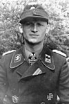 A black-and-white photograph of a man wearing a military uniform, cap, and a neck order in shape of an Iron Cross. His cap has an emblem in shape of a human skull and crossed bones.