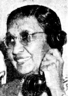 A South Asian woman, smiling, wearing glasses, with a phone to her ear.