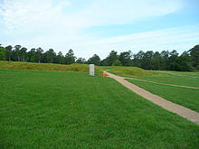 A wide grassy field with a paved walkway running through it. In the distance is a small stone marker.