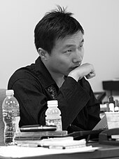 Black-and-white photo of an Asian man seated at a desk, holding his fist to his chin.