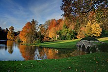 Farbfotografie eines Sees mit einer kleinen Bogenbrücke aus Stein am rechten Bildrand. Im Hintergrund eine Wiese und Herbstbäume. Im Vordergrund ist ebenfalls eine Wiese.