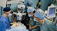 A surgical team is gathered around the patient in an operating theatre. the surgeon and two learners are observing the procedure through a surgical microscope suspended above the patient's eye.