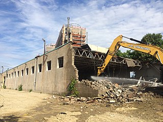 A cinderblock building being demolished by an excavator