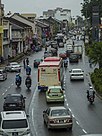 A city street lined with shophouses, with cars, lorries, motorcycles and a bus on the road.