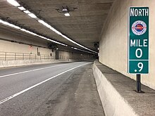 A two-lane highway running through a tunnel. A green mile marker sign in the foreground reads NORTH / MILE 0.9