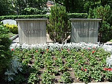 Frontale Farbfotografie von zwei einfachen Grabsteinen mit Nadelsträuchern neben ihnen. Auf dem linken Stein steht „Oona Chaplin 1925-1991“ und auf dem rechten Stein steht „Charles Chaplin 1889-1977“. Im Vordergrund sind in der Erde grüne und weiße Topfpflanzen. Im Hintergrund sind Gräber und eine Hecke.