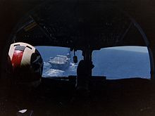 Photograph of the interior of an aircraft cockpit, taken from behind the pilot, who is sitting on the left hand side. An aircraft carrier is visible through the left-hand pane of the cockpit windows.