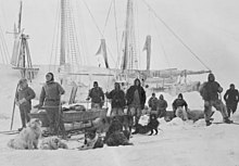 A group of men pose on the ice with dogs and sledges, with the ship's outline visible in the background