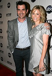 A blonde woman wearing a silver dress smiles as she stands on a red carpet to the left of a man with dark hair wearing a blue striped shirt, gray blazer, and black pants. They are both standing in front of a backdrop with "NYTVF" and a logo that is a round black ball with the letters "abc" on it