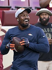 Von Miller wearing a cap and AirPods smiles while holding a football. He is also wearing gloves and looks like he might soon throw the ball.