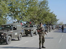 A soldier stand in front of a convoy of military vehicles, with KFOR markings and Portuguese flags. The second véhicule is a Portuguese VBL.