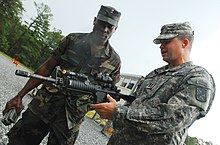 A colour photograph of two soldiers, one holding an assault rifle, talking at a training ground. The soldier on the left is in a camouflage field uniform, while the soldier on the right holding the rifle is in a grey camouflage uniform