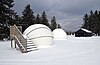 Two white observatory domes in a snow-covered field, wooden stairs are beside one dome and a cabin and trees are in the background