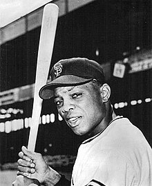 Photograph of Mays with SF on his black cap staring towards the camera holding a bat on his right side, with a baseball grandstand in the background