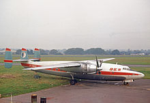 Twin-engined, triple-tailed, high-winged propeller-driven passenger aircraft, in profile on an airport taxiway. Natural metal finish except for airline insignia and a lengthwise stripe along the length of the fuselage.