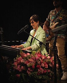 Young woman singing into a microphone with a big smile while playing the piano