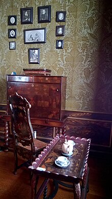 A photograph of a room decorated in green wallpaper, with several pieces of wooden furniture as well as an arrangement of miniature black and white portraits