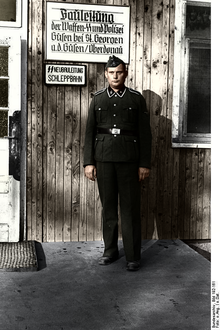 SS-Mann in Uniform vor hölzerner Baracke mit Schild „Bauleitung“