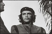 Black-and-white image of a man looking to his right. He wears a black beret with a little "x" and a closed jacket. His hair is shoulder-length, curly and black. On his right there is a man looking to him and on his left, there is a plant.