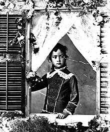 A young Princess Kaiulani, standing, framed by a window with left hand resting on the window sill.