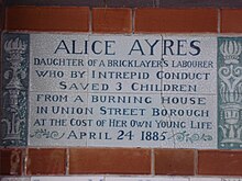 A tablet formed of six standard sized tiles, bordered by green flowers in the style of the Arts and Crafts movement, with an inscription in green capital letters on a white background