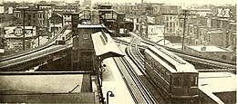 A one-car train with arched windows waits at the junction while a wooden car is at the southbound "L" platform. The image is sepia looking eastward, a similar view to the introductory image.
