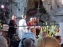Seitliche Farbaufnahme vom Papst in rotgoldener liturgischer Kleidung, der mit vier Würdenträgern vor einem Altar steht und seine Hände zum Gebet zusammenhält. Die Gruppe ist gut beleuchtet und Kameras befinden sich vor dem Podium.