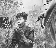 A scarred and bloodied man of Oriental appearance kneels on the ground. Beside him stands a man with an FN FAL battle rifle. A bayonet is fixed to the end of the rifle.