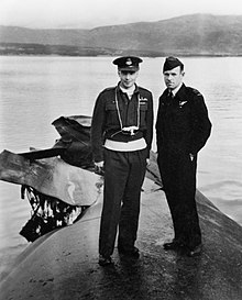 Black and white photo of two men wearing military uniforms standing on the overturned hull of a ship