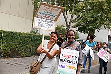 Zwei Schwarze Frauen halten Poster, auf einem steht „Generation Adefra. Schwarze Feministinnen* sagen NEIN zu Rassismus, Frauenhass, Homo-/Transphobie, Trumpismus, Faschismus!“. Auf dem anderen Plakat steht: „I love my Blackness and yours“.