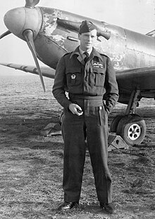 A full length picture of a man in RAF uniform. He stands in front of a parked aircraft, with one hand in a pocket and holding a pipe in the other.