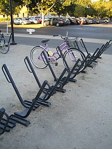 An array of sixteen or so parking spots for bicycles. Each is a square metal bar angled at 45 degrees from the ground with a parallel loop of thinner round tubing at the top. They are set in a row with the bars pointing in alternating directions. A pink ladies' bicycle with a shipping basket is parked in one. Another row of them is mostly out of frame to the left with a mountain bike parked in it. In the background is a car park with trees.