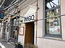 Altes Haus in Nahaufnahme mit einem Gerüst für Fassadenrenovierung davor. Über dem Eingangstor in der Mitte ist ein Schild mit der Aufrschrift „Inigo“.