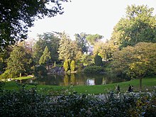 Farbfotografie eines runden Weihers, der von Wiesen und Bäumen umgeben ist. Links führt ein Wasserzulauf zu einer tieferen Ebene. Im Vordergrund verdecken Sträucher mit kleinen Blüten teilweise den Weg mit drei Menschen.