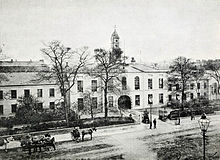 A black and white photograph, showing a stately-looking two-storey building with white walls, extending out of shot to the left and right, with an arched cart entrance at the centre. A modest clocktower rises above the entrance, and the building is surrounded by neat shrubbery and iron railings. A wide street crosses left-right outside of the fence, with a handful of horse-drawn carts and pedestrians in 19th-century clothing.