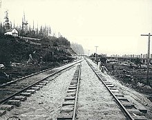 A black-and-white photograph of two railroad tracks against an undeveloped hillside with trees and a small house.