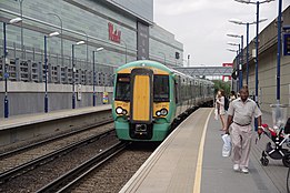A green Southern Railways train is stopped at a platform. In the background is a silver building. A sign on the building reads, "Westfield".