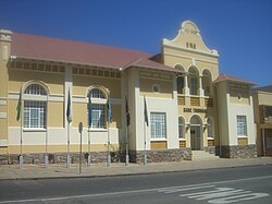 A yellow and white neo-classicist building in bright sunlight. The roof of corrugated iron sheets is painted dark red, on the main gable the inscriptions read "A.D.1913" and "SADC Tribunal". Seven flagpoles with national flags of countries from the Southern African Development Community are mounted onto the pavement in front of the building.