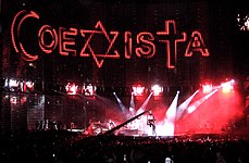 A view of the stage being filmed, with "COEXISTA" spelled out on the video screen, using an Islmaic crescent as a "C", a Star of David as an "X", and a Christian cross at a "T". A camera in the middle shows the concert being filmed.
