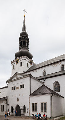 Frontale Farbfotografie von einer weißen Kirche mit schwarzer Dachhaube. Der Rundbogeneingang befindet sich in einem Vorbau mit Giebelfeld. Die Fassade ist schlicht gehalten. Im Vordergrund sitzen Menschen auf einer Bank.