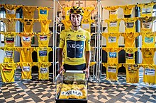 Egan Bernal wearing his team kit and yellow jersey standing in front of a museum exhibition of historic yellow jerseys