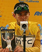 Cavendish sitting in front of a microphone holding a copy of his book Boy Racer