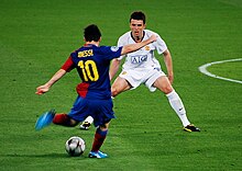 A man wearing a blue-and-red football shirt prepares to kick a football, while a man in a white shirt looks on.