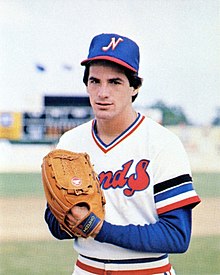 A man wearing a white baseball jersey with long blue sleeves beneath and a blue cap with a white "N" on the center has his hands bought together in his glove and a determined look on his face.