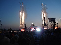 A large crowd is gathered around a stage, with fireworks in the sky