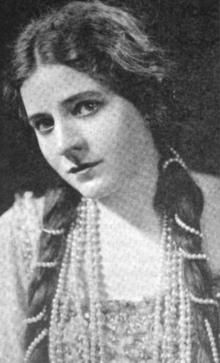 A young white woman wearing her long dark hair parted center and in long braids wrapped in pearls, with further long strands of pearls around her neck